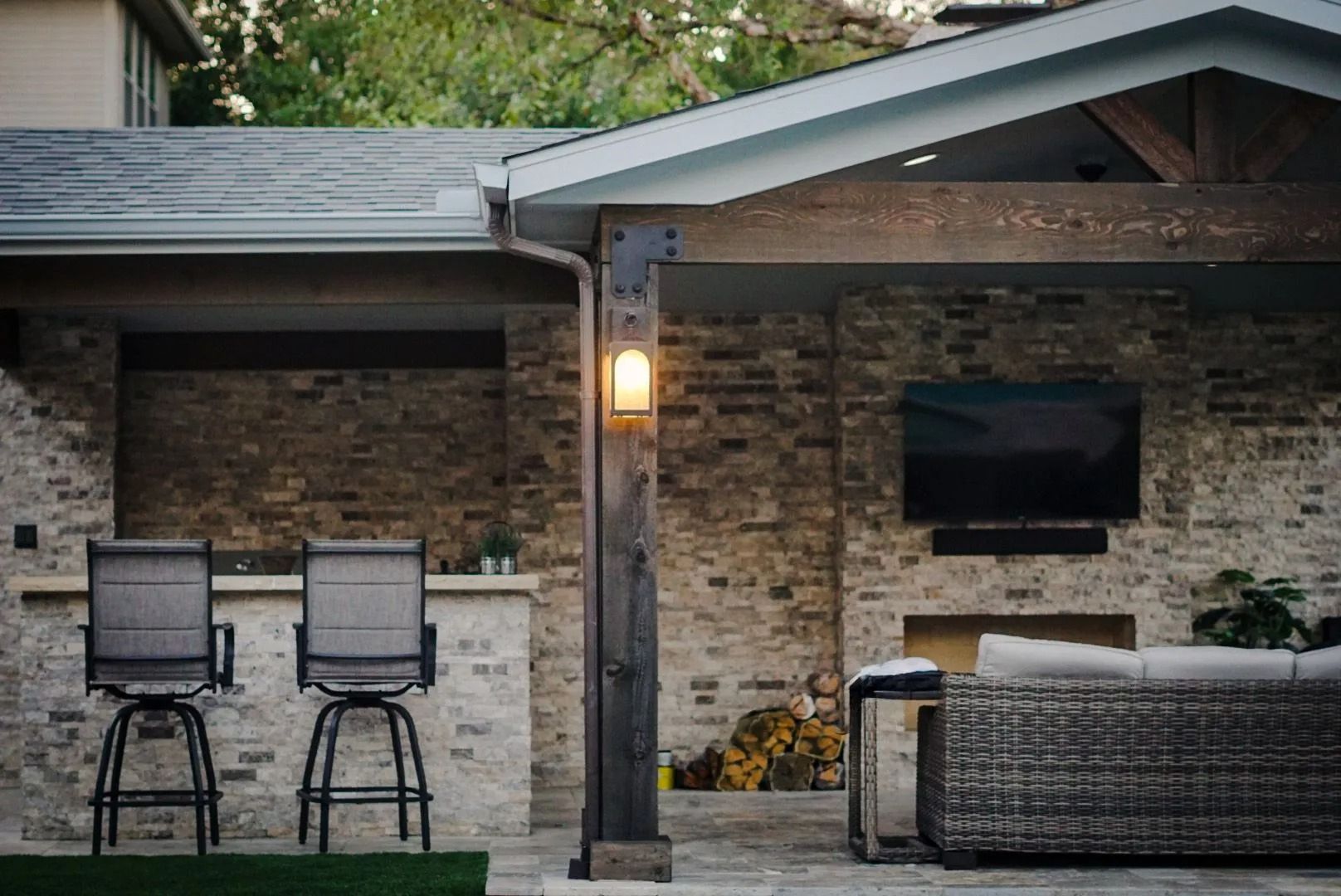 Covered brick patio with two stools, wall-mounted TV, glowing lantern, and wicker sofa area