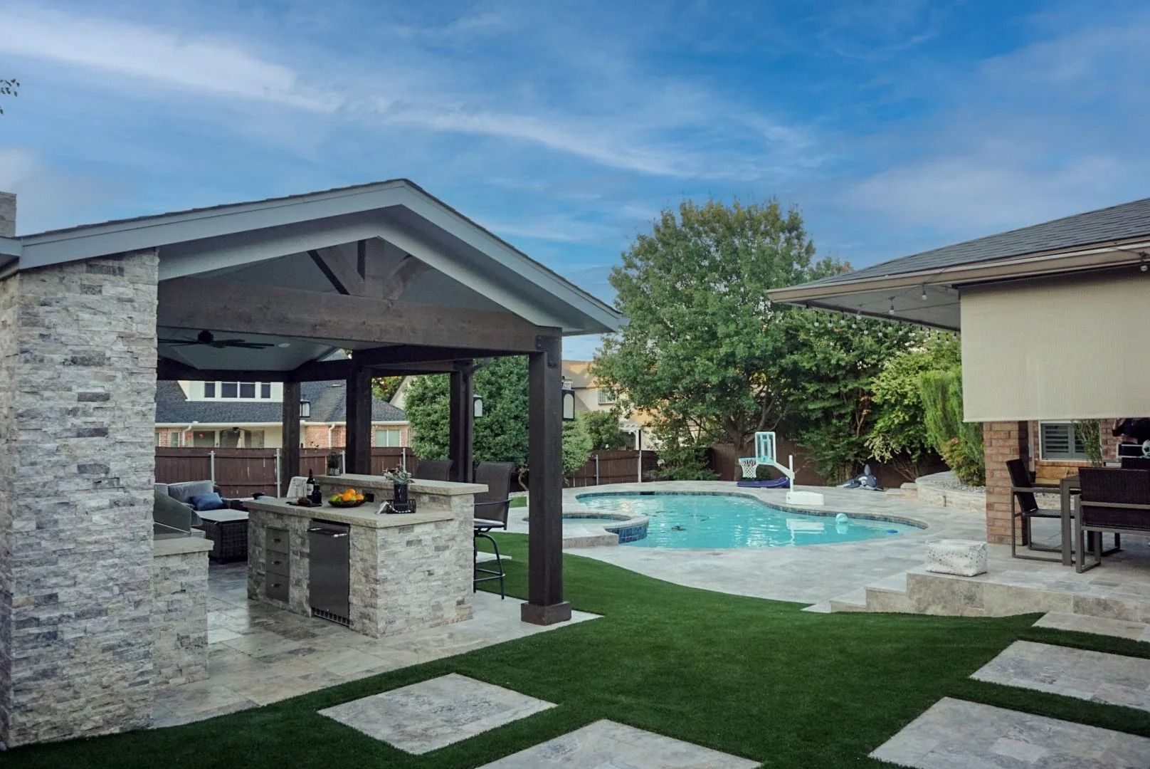 Backyard patio with pool, stone pavilion, and green lawn under a cloudy sky