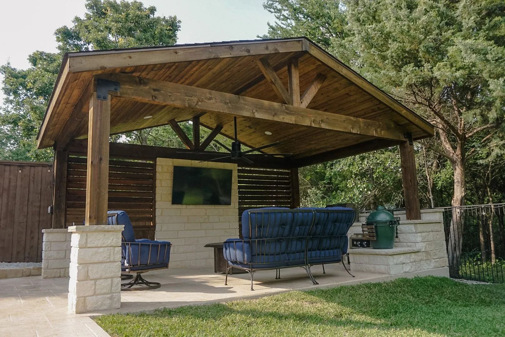 Wooden backyard pavilion with cushioned outdoor seating on a patio, surrounded by trees and grass.