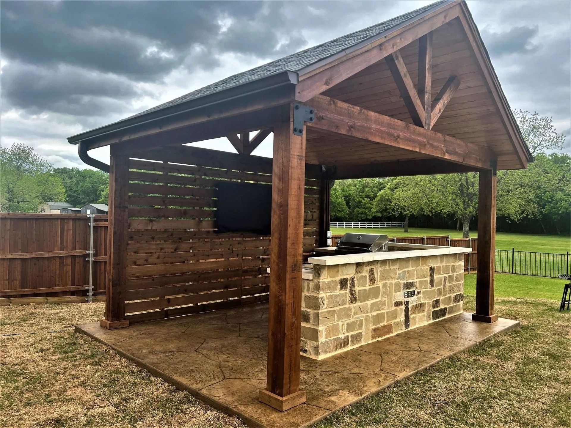 Wooden outdoor pavilion with a stone bar counter on a paved patio, beside a fenced grassy yard.