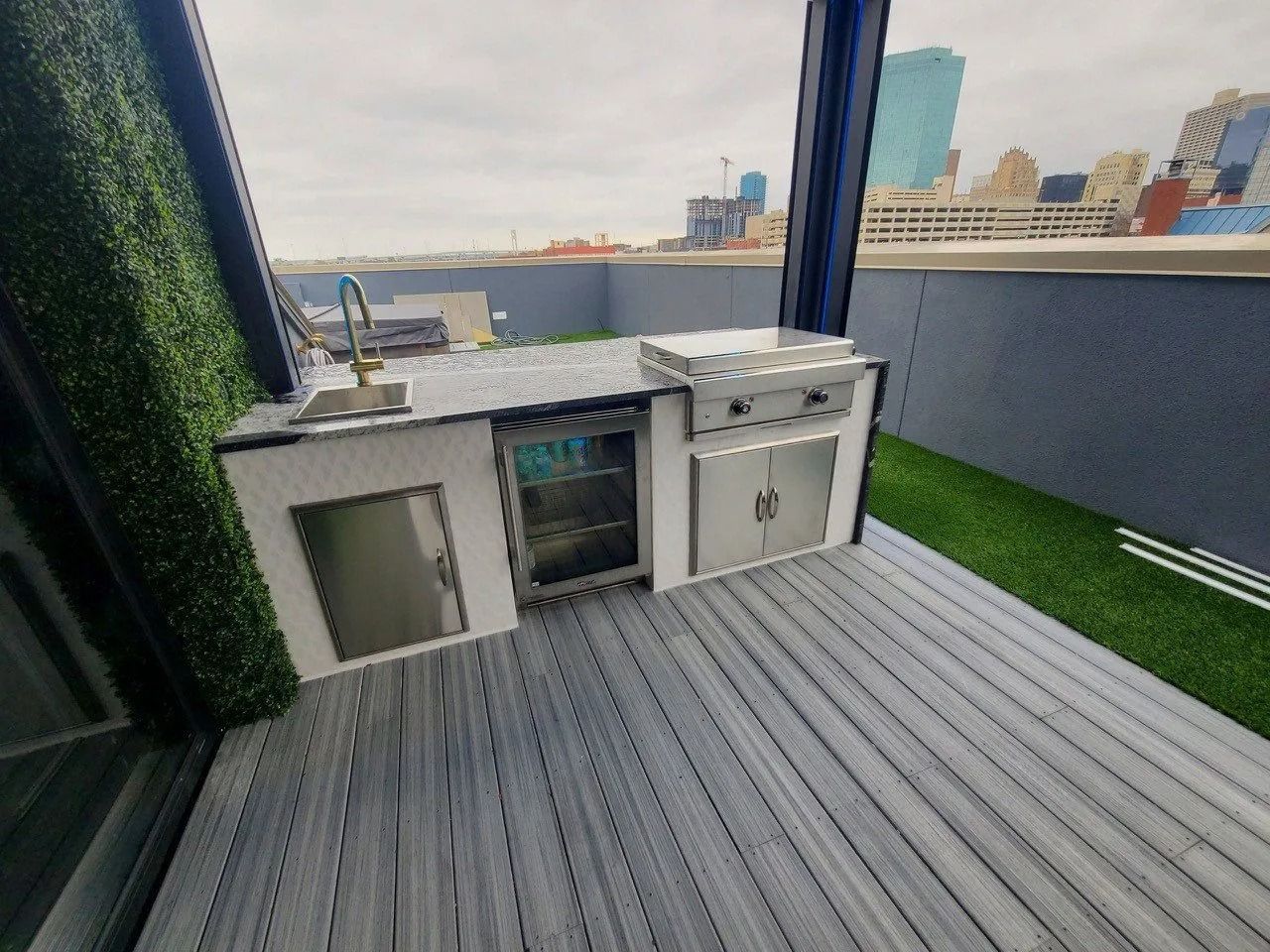 Outdoor rooftop bar with sink, mini fridge, and grill on gray decking overlooking the city skyline