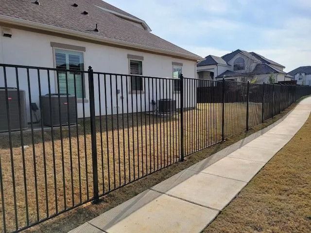 Black metal fence along a concrete sidewalk beside a suburban house and dry yard.