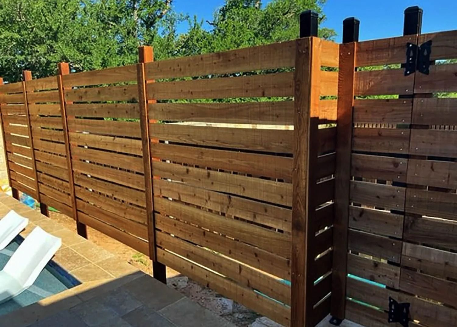 Brown wooden privacy fence along a patio, with trees and blue sky in the background.