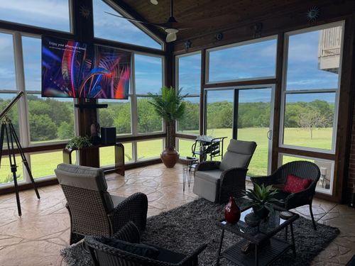 Screened porch seating area with TV, chairs, table, and a view of green fields beyond large windows
