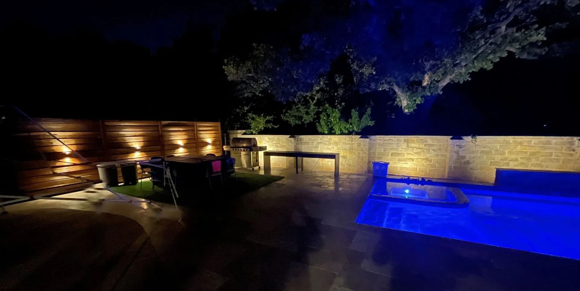 Nighttime backyard patio with glowing landscape lights and a blue-lit pool or hot tub.