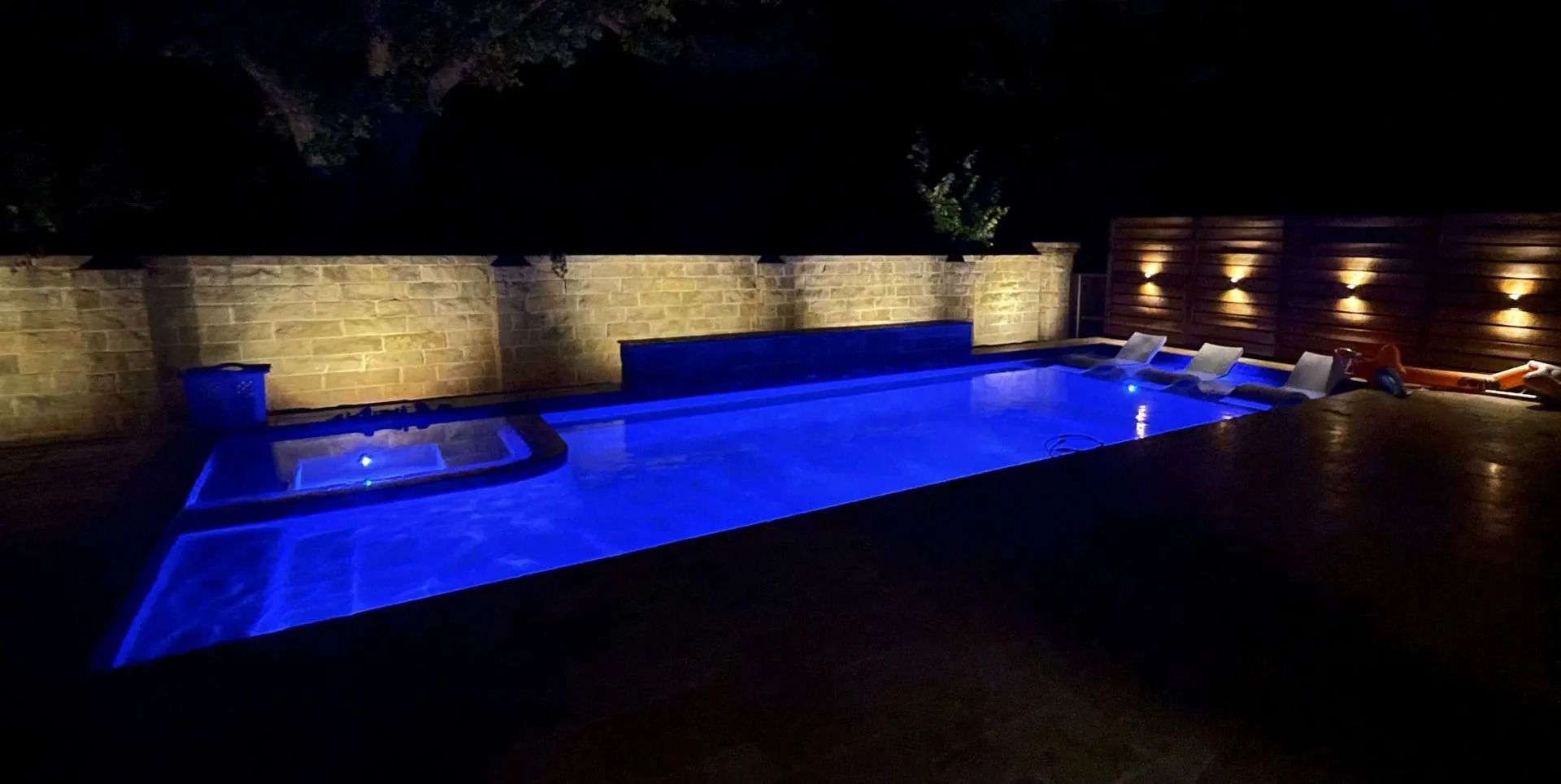 Blue-lit outdoor pool beside a stone wall at night, with lounge chairs along the deck