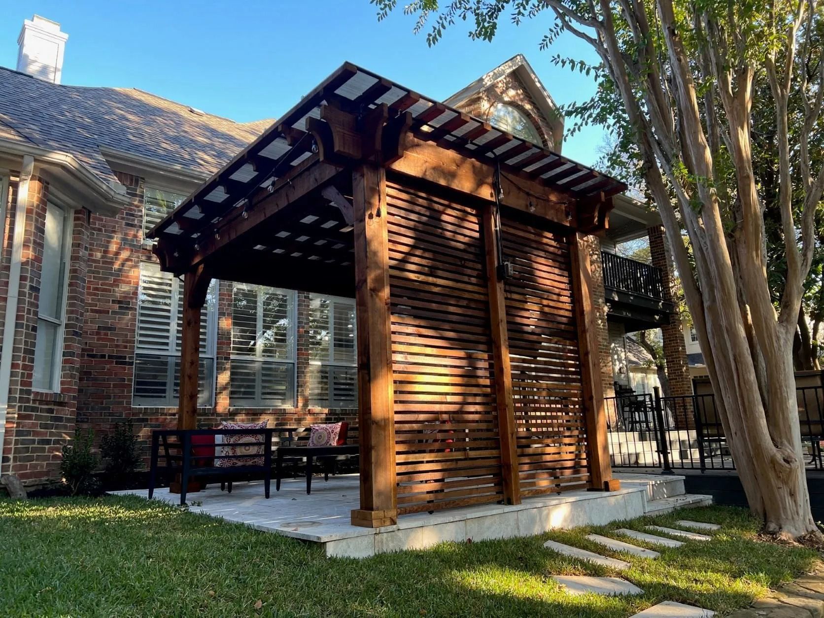 Wooden patio pavilion beside a brick house, with slatted walls and trees in the yard.