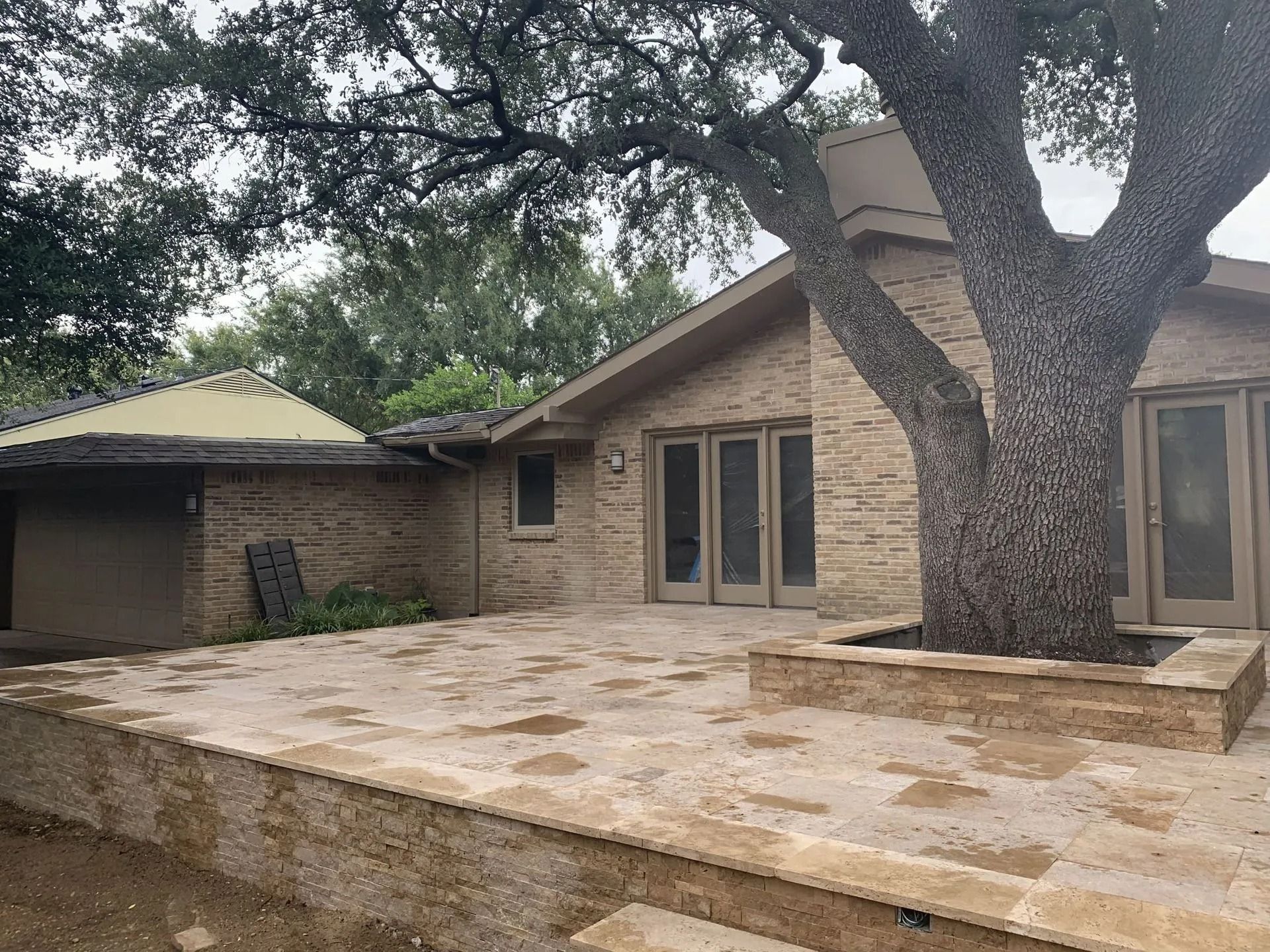 Stone patio and large tree in a backyard beside a beige brick house with a covered porch.