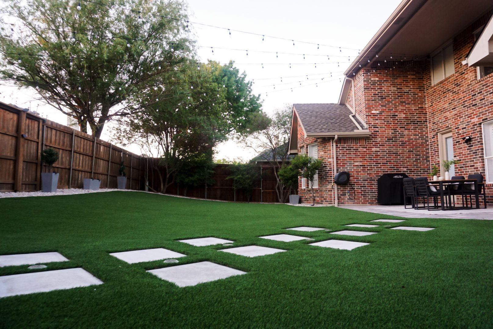 Backyard patio with artificial turf, stepping stones, and brick house at dusk
