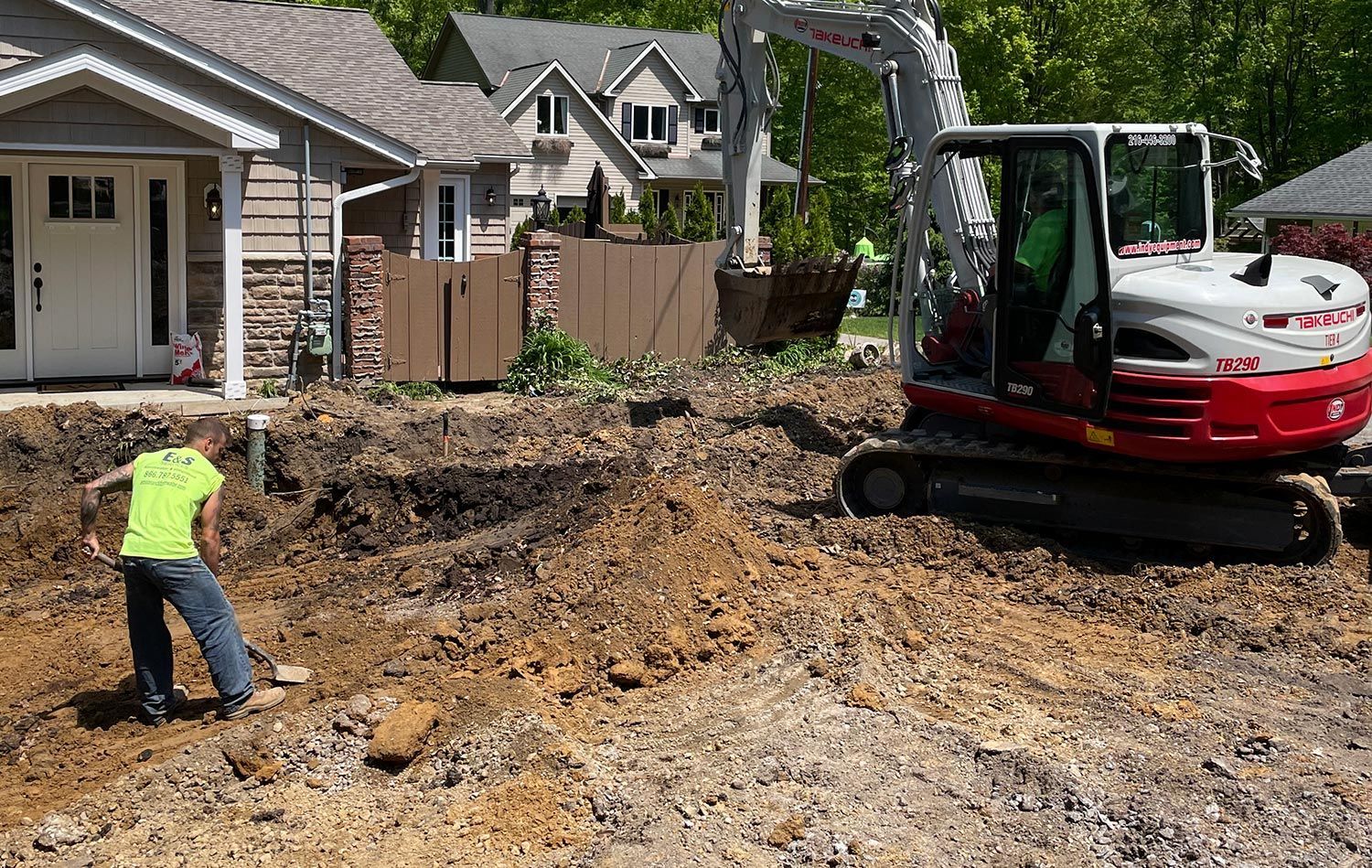 A man is digging in the dirt in front of a house with an excavator in the background.