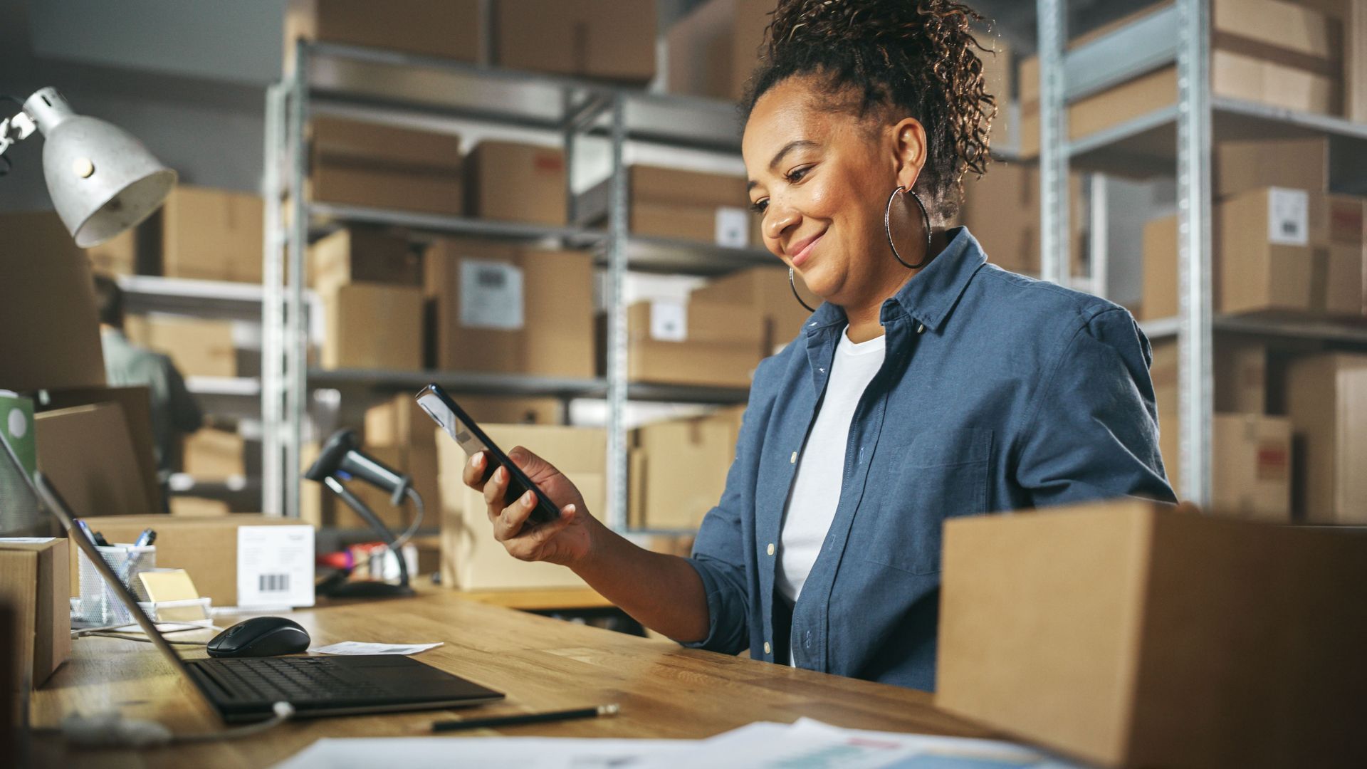 Woman in blue shirt smiles at phone, surrounded by boxes in a warehouse setting.