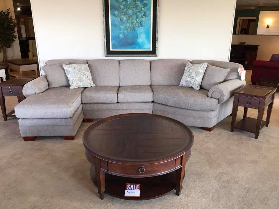 Beige sectional sofa with a round wooden coffee table and two end tables in a furniture store.