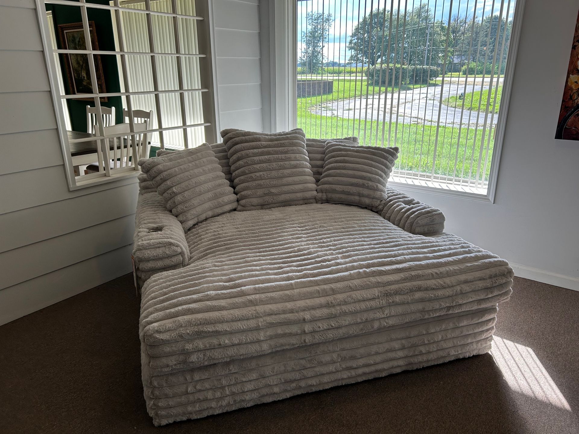 Cozy, light gray, plush chaise lounge with three pillows near a window with vertical blinds.