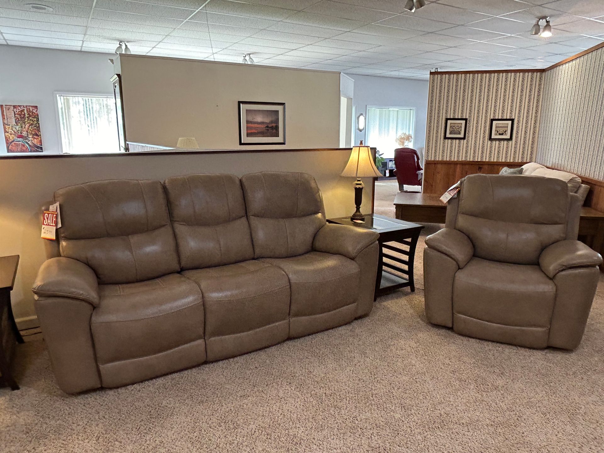 Tan leather sofa and recliner in a carpeted showroom setting. A lamp and table are nearby.