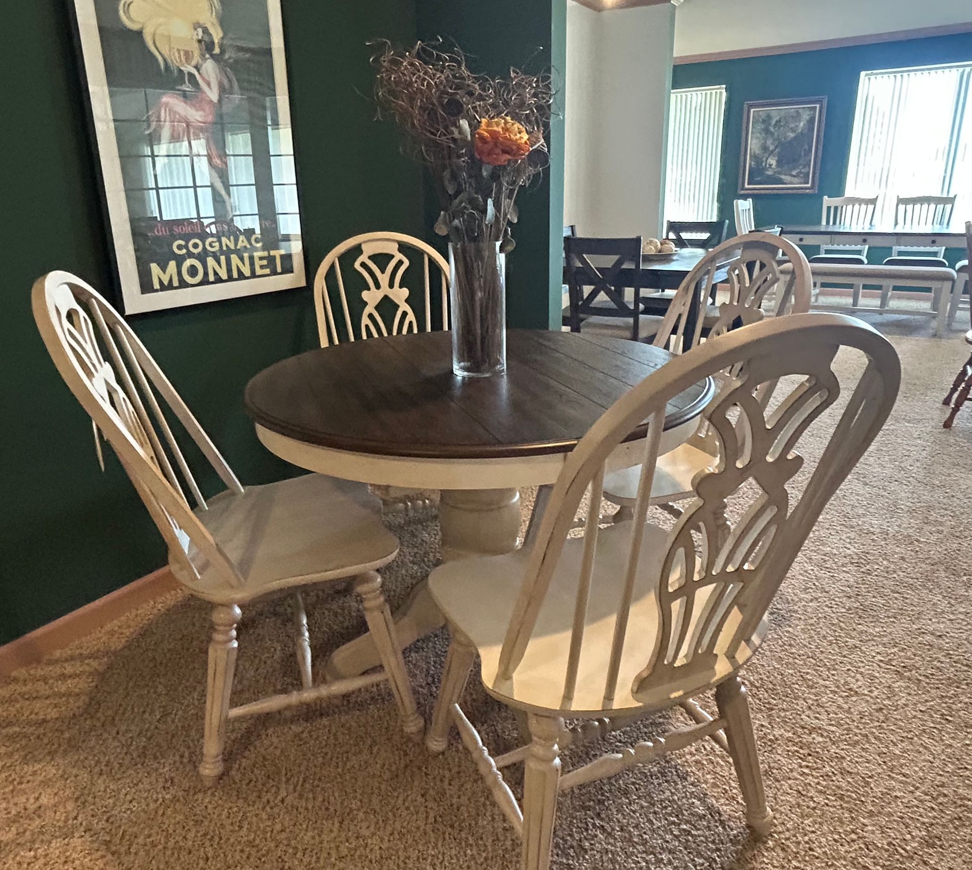 Round wooden dining table with four white chairs; poster, and flower vase against green wall.