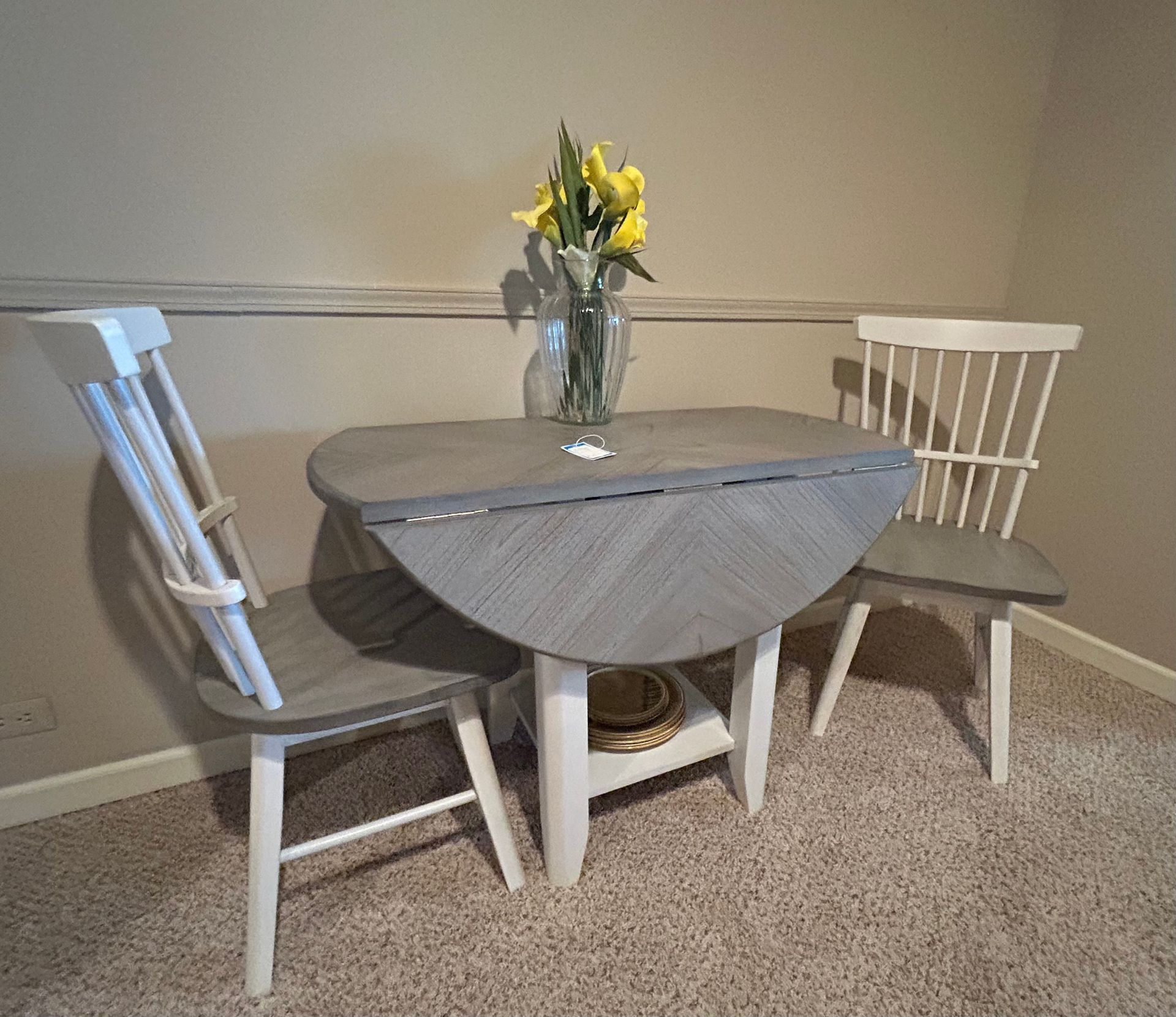 Small, gray drop-leaf table with two white chairs, vase of yellow flowers. Set against a beige wall, on carpet.