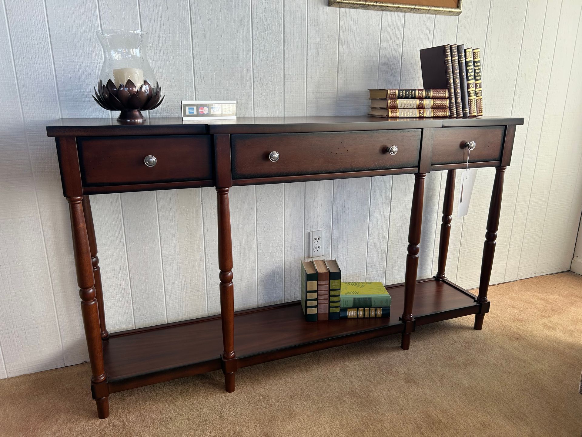 Brown console table with three drawers, books, and a candle holder against a white paneled wall.