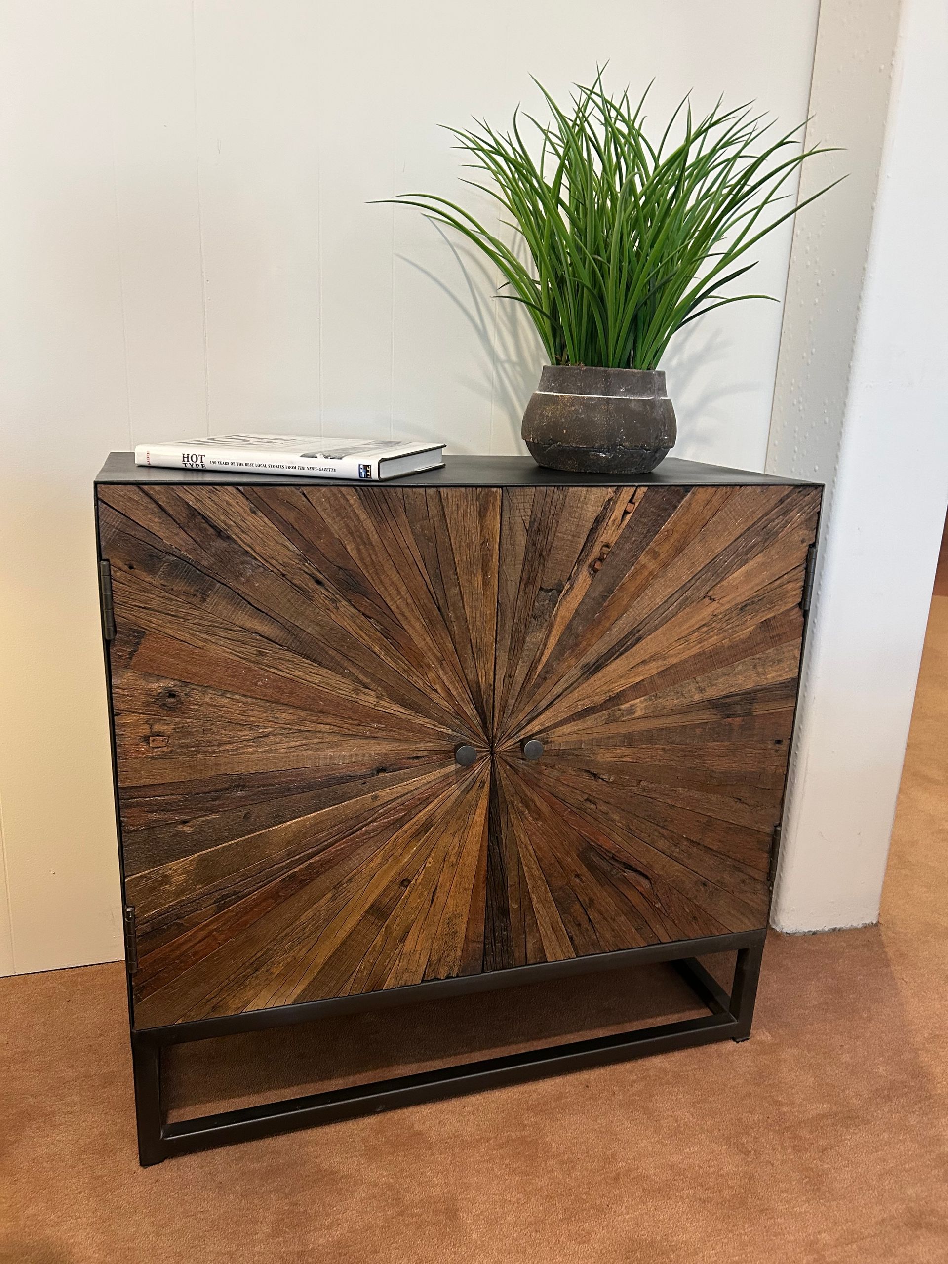 Wooden cabinet with sunburst design, topped with a plant and books; brown and dark tones.