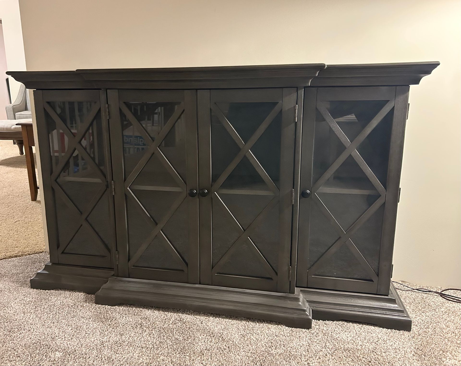 Dark gray wooden cabinet with four glass-fronted doors, cross-shaped accents, and a wide base, in a room with carpet.