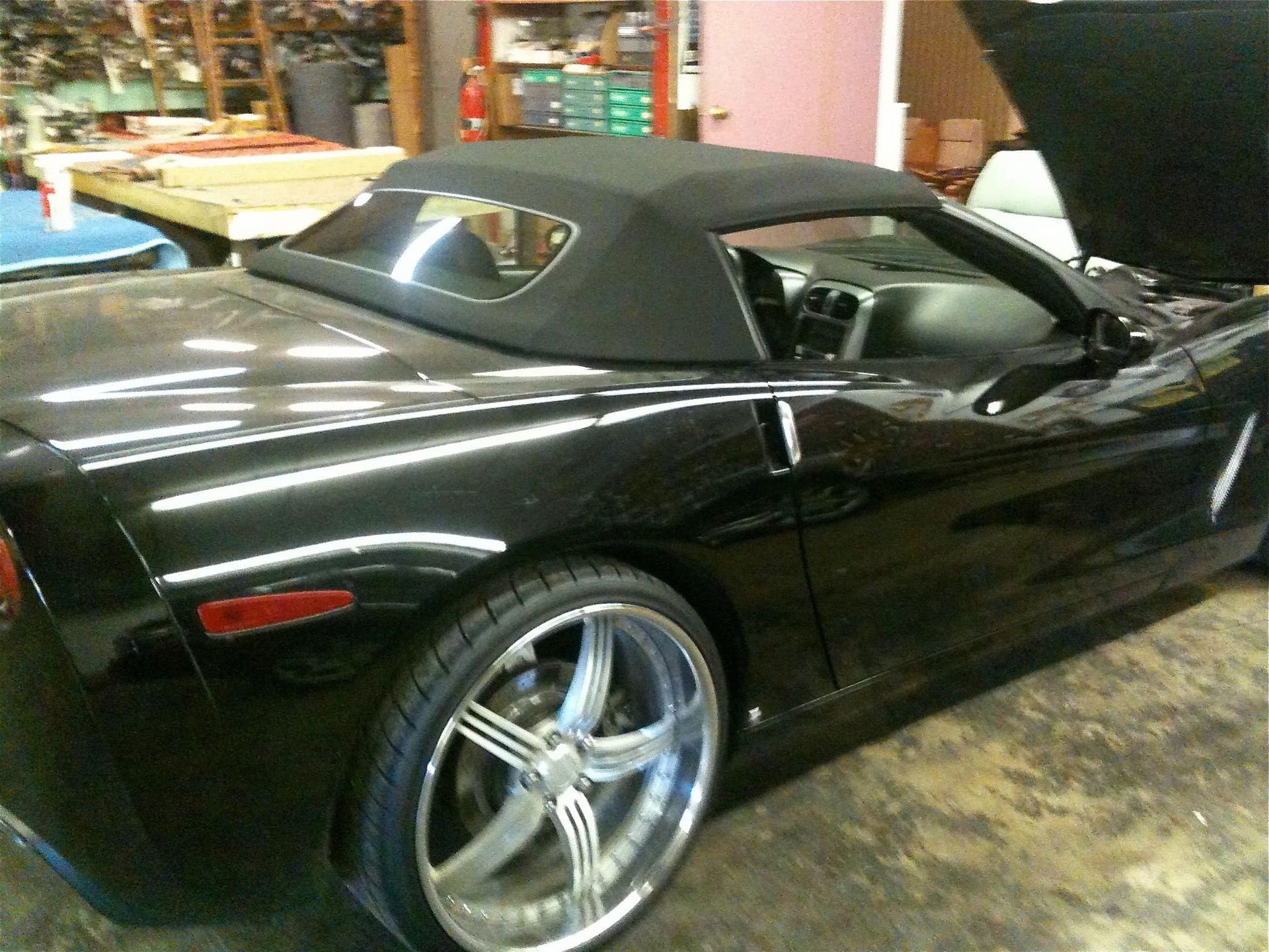 Black convertible sports car with top up, chrome wheels, inside a shop.
