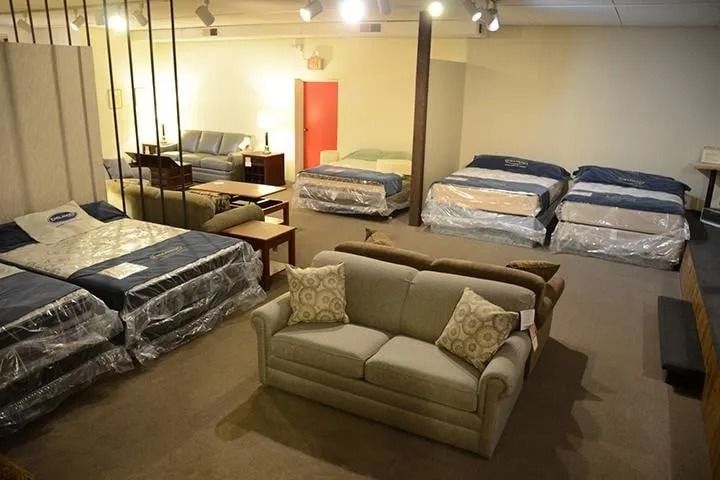 Furniture showroom with beds, sofas, and tables. Some items are wrapped in plastic, beige and wood tones.