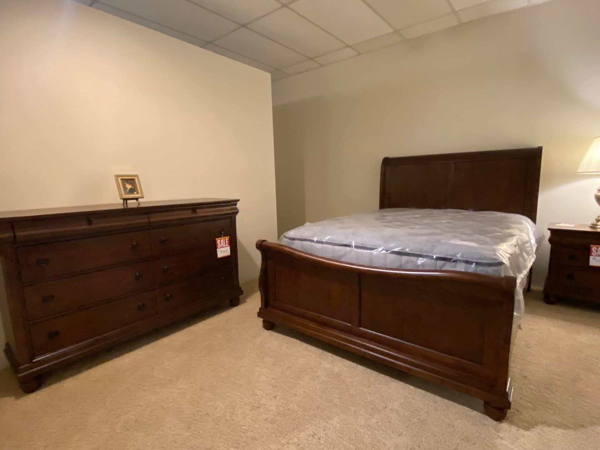 Bedroom furniture set: dark brown bed frame, dresser, nightstand, neutral wall and carpet.