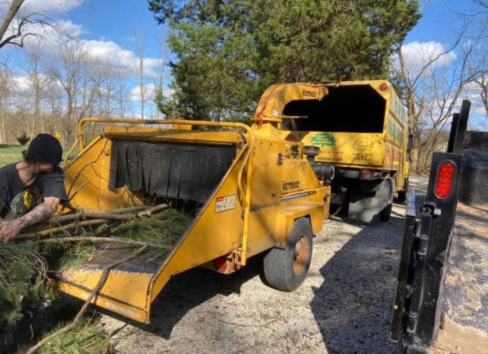 Yellow wood chipper and dump truck in a gravel yard, with a worker loading branches into the machine.