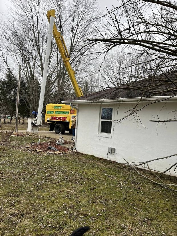 Yellow utility bucket truck working beside a white house, lifting near a roofline in a yard with bare trees