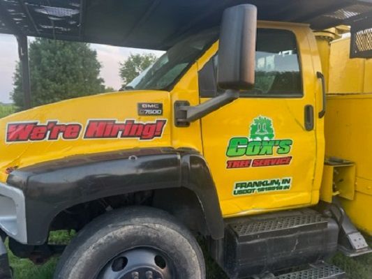 Yellow work truck with “We’re Hiring!” sign and company logo under a canopy