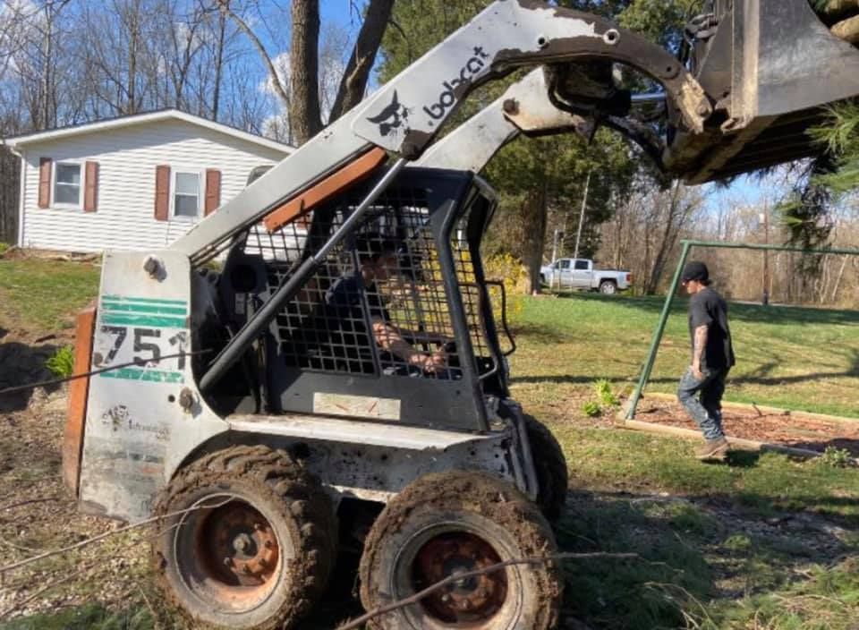 Skid-steer loader with raised bucket lifting tree limbs beside a house, with a person walking nearby