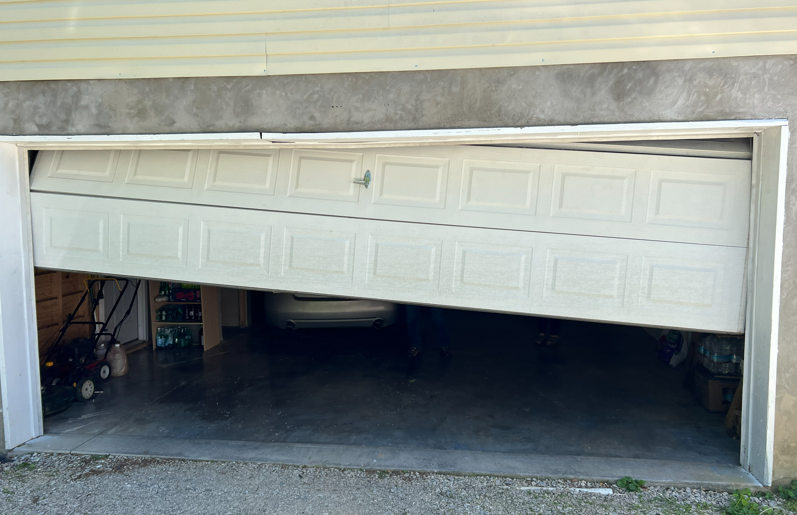 A white garage door is stuck halfway open and crooked, with its left side tilted significantly lower than the right side.