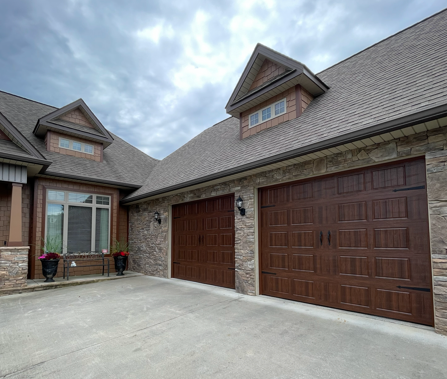 A stone-facade house exterior with two brown garage doors and two gabled windows under a cloudy sky.