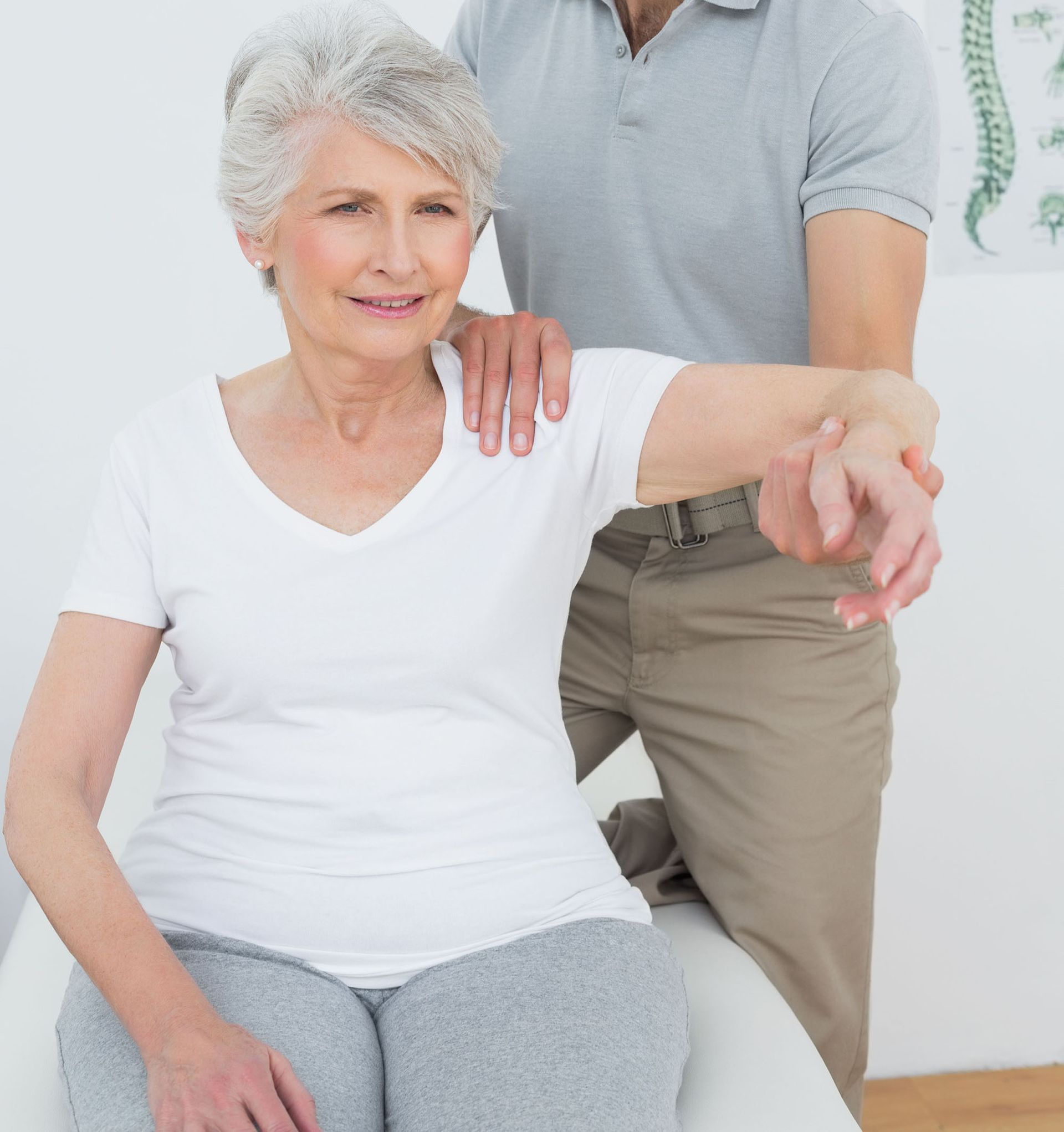 Older woman doing a shoulder stretch with a caregiver supporting her arm in a bright room