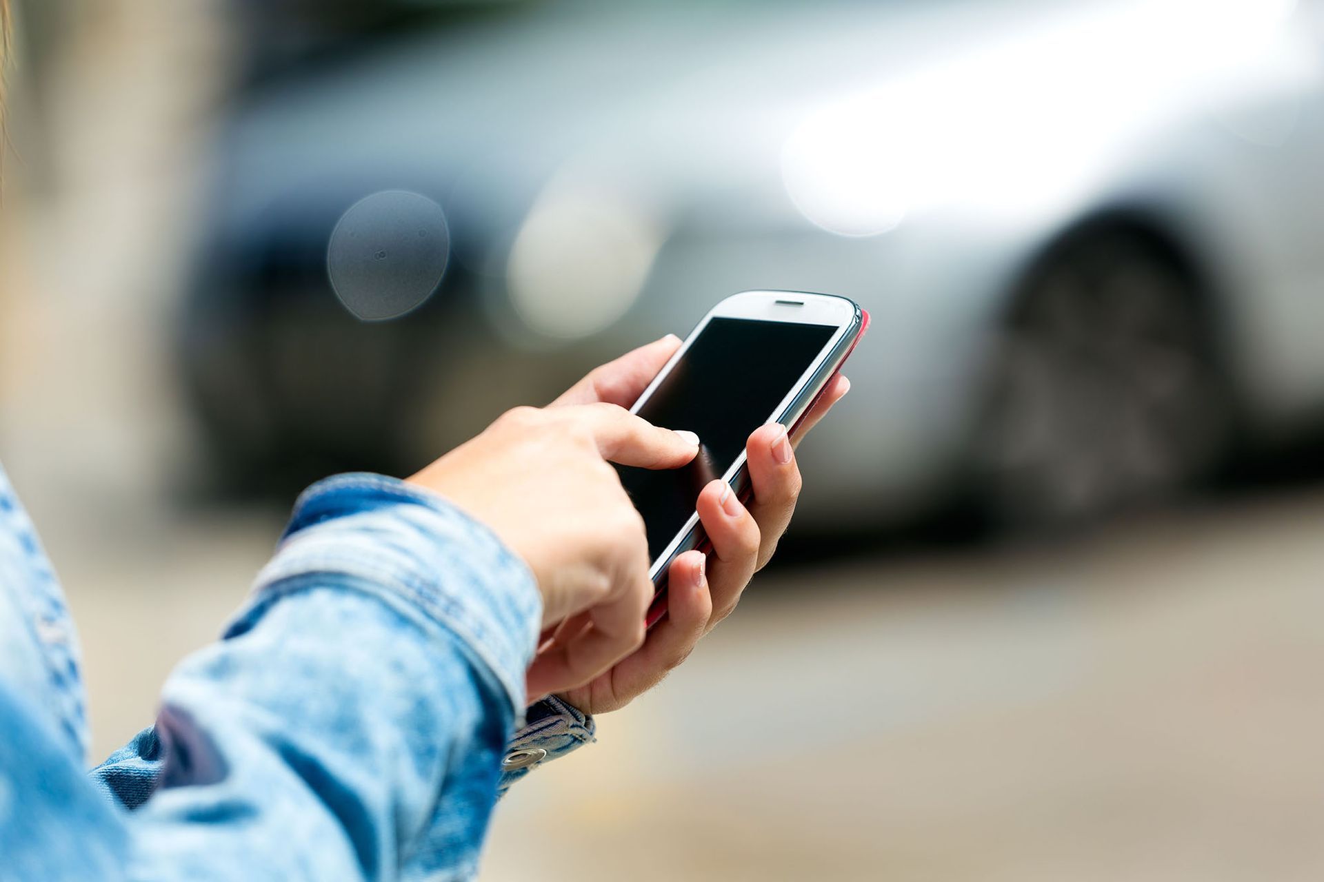 Hand holding a smartphone outdoors with a blurred car in the background