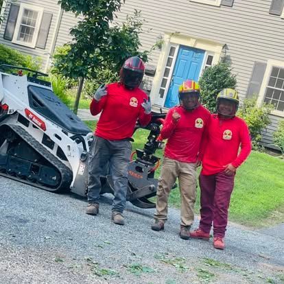 Three workers in red shirts and helmets pose next to a Bobcat, in front of a house.