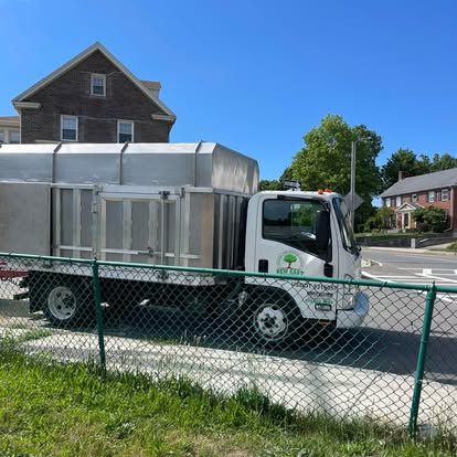 White truck with a silver container on the back parked on a street near a green fence, houses, and trees.