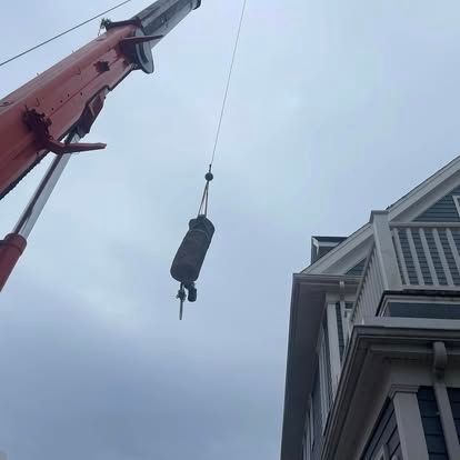 Crane lifting a cylindrical object near a blue house on a cloudy day.