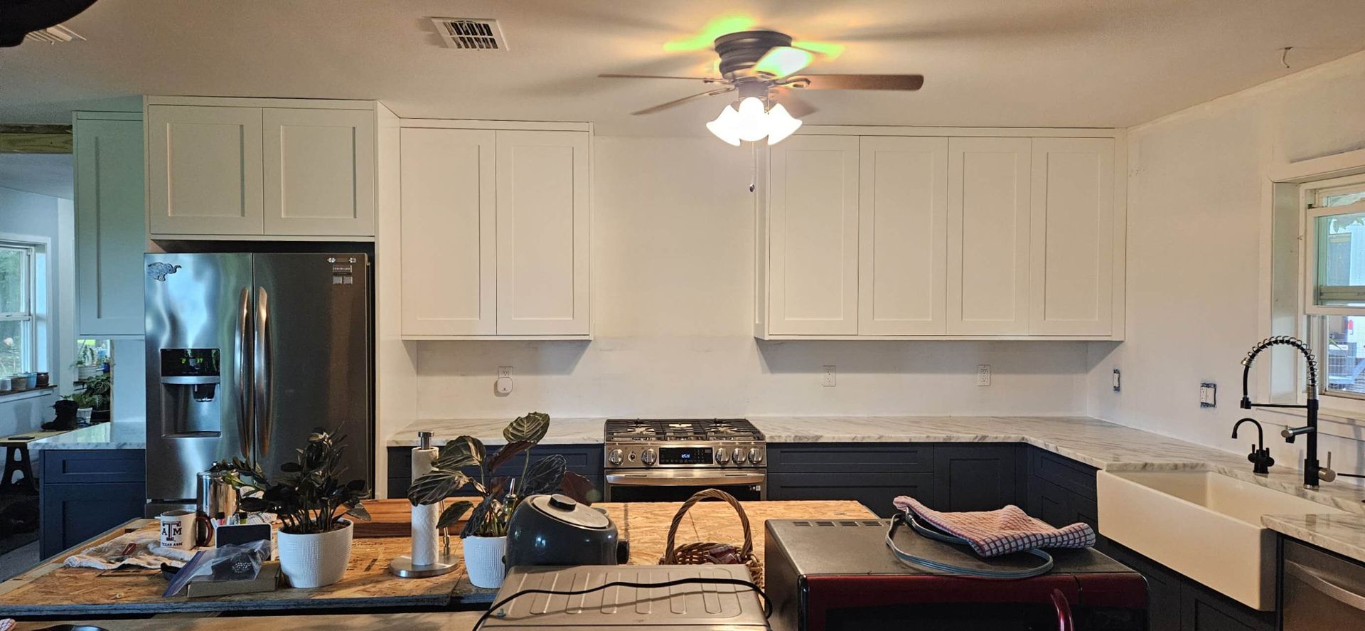 A kitchen with white cabinets , a stove , a refrigerator , a sink and a ceiling fan.