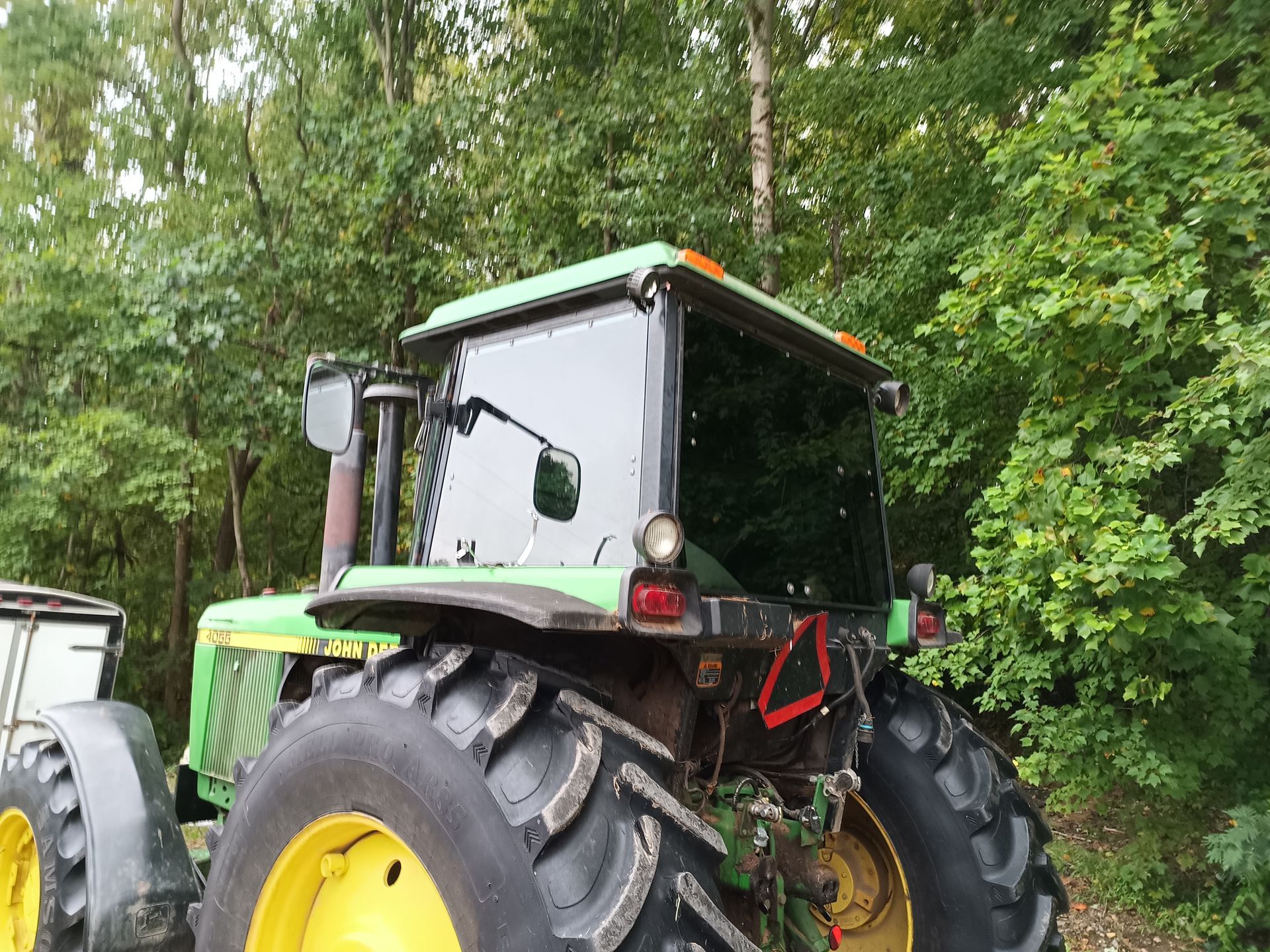 John Deere tractor, green and yellow, in a wooded area. Rear view with large tires and enclosed cab.