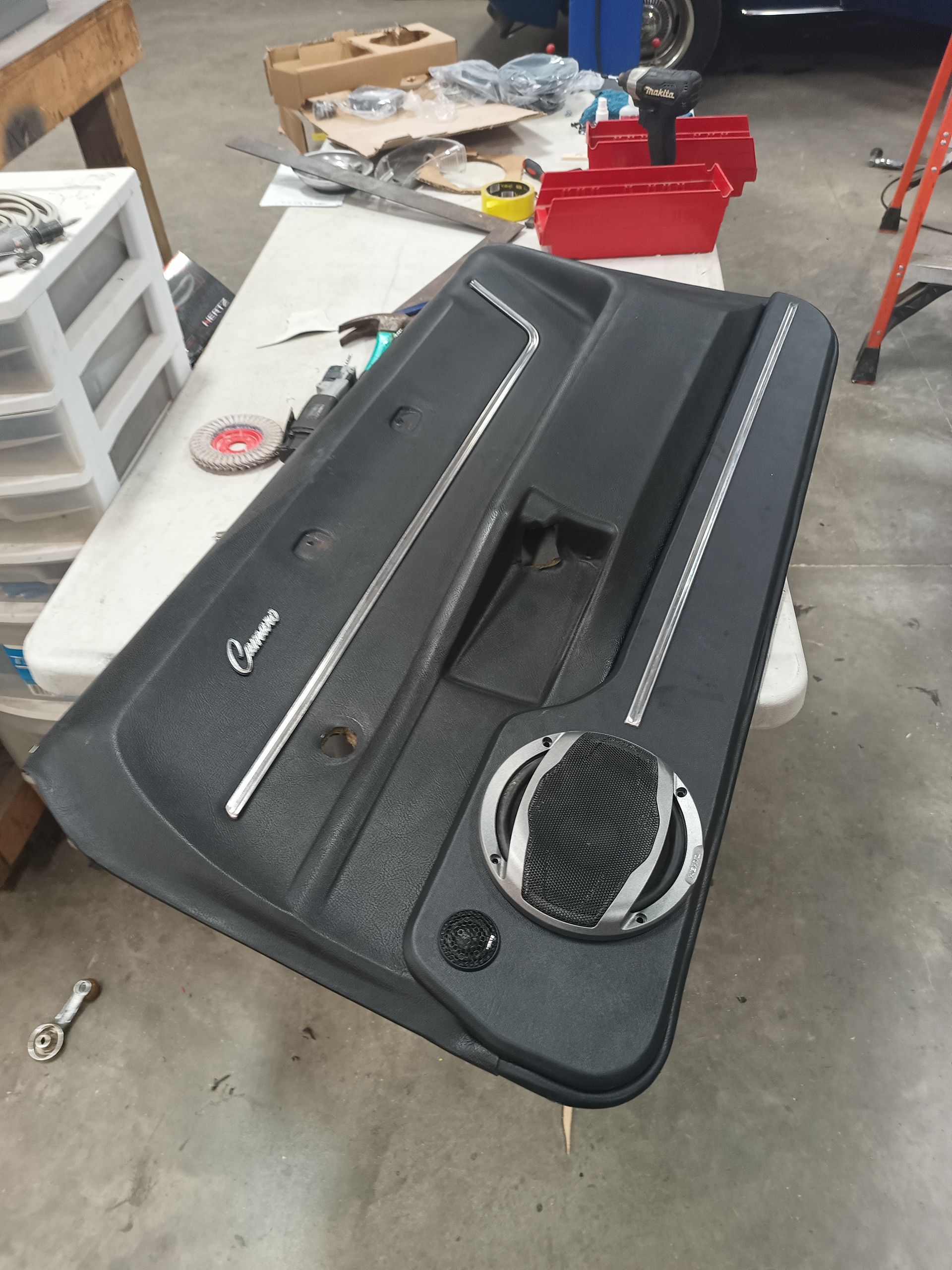 Black car door panel with speaker, silver trim, on a table in a garage.