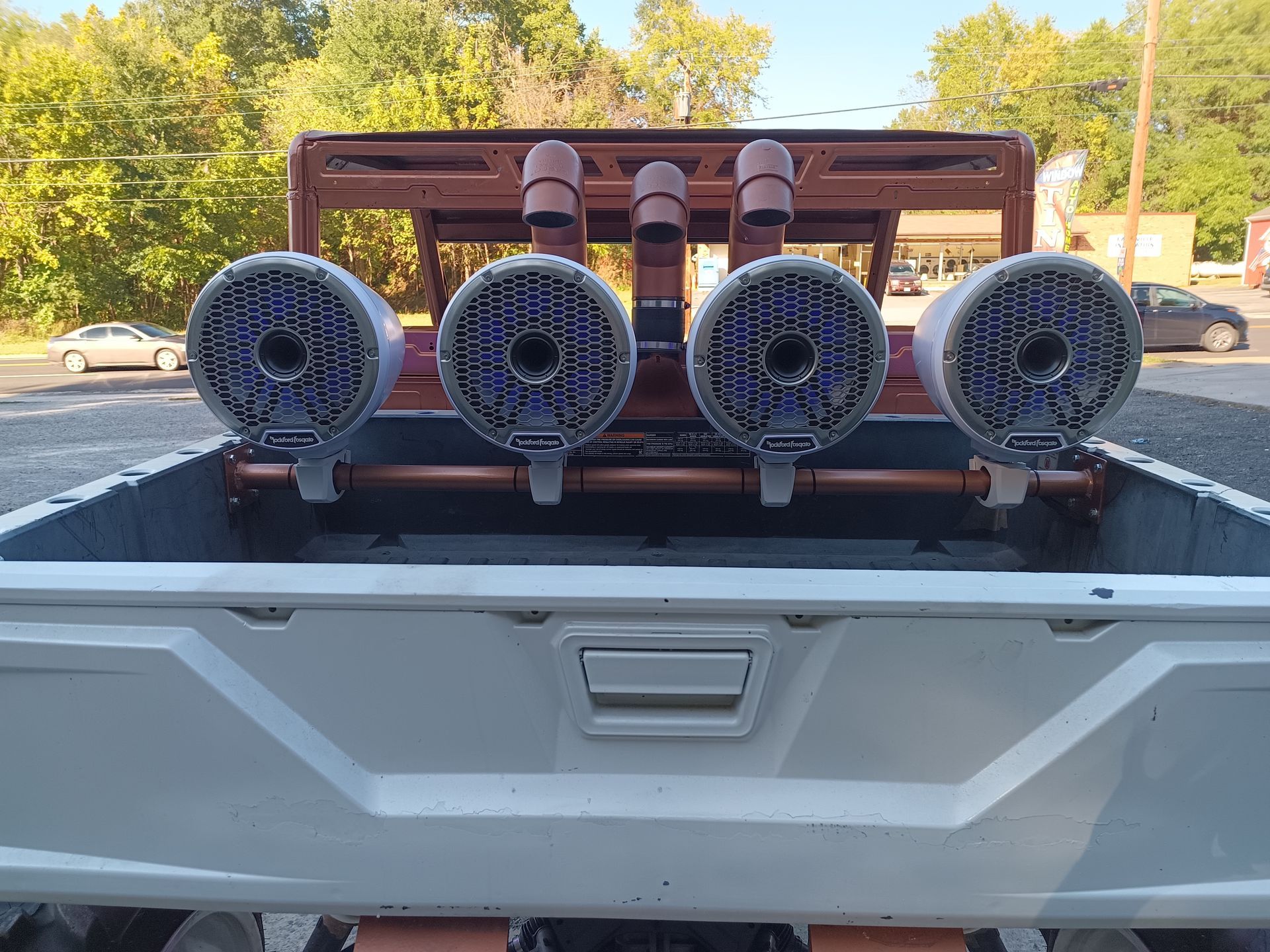 Back of a white pickup truck with four large speakers mounted on a copper frame.