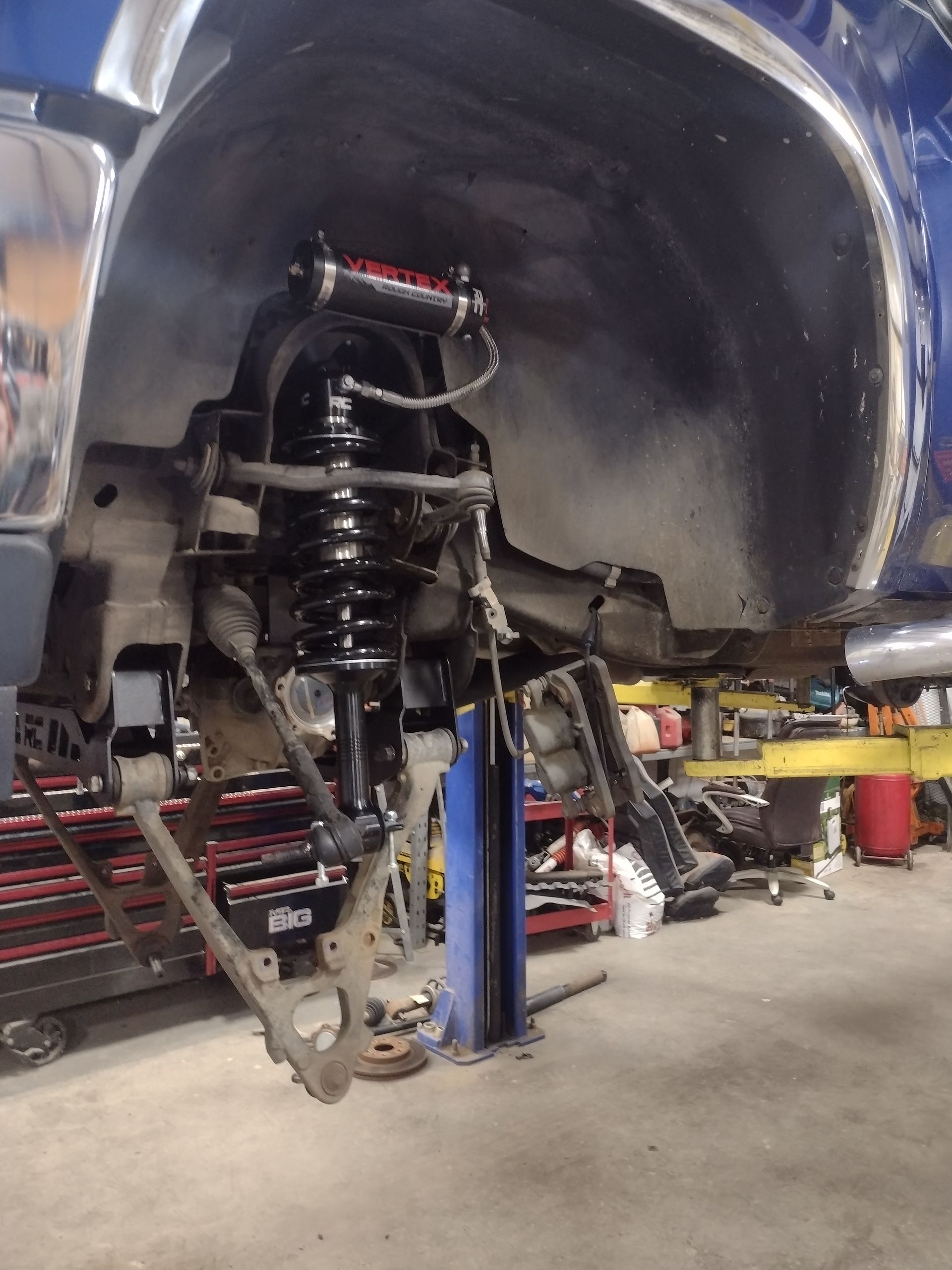 Blue truck's front suspension being worked on in a shop; a lifted wheel well and exposed components are visible.