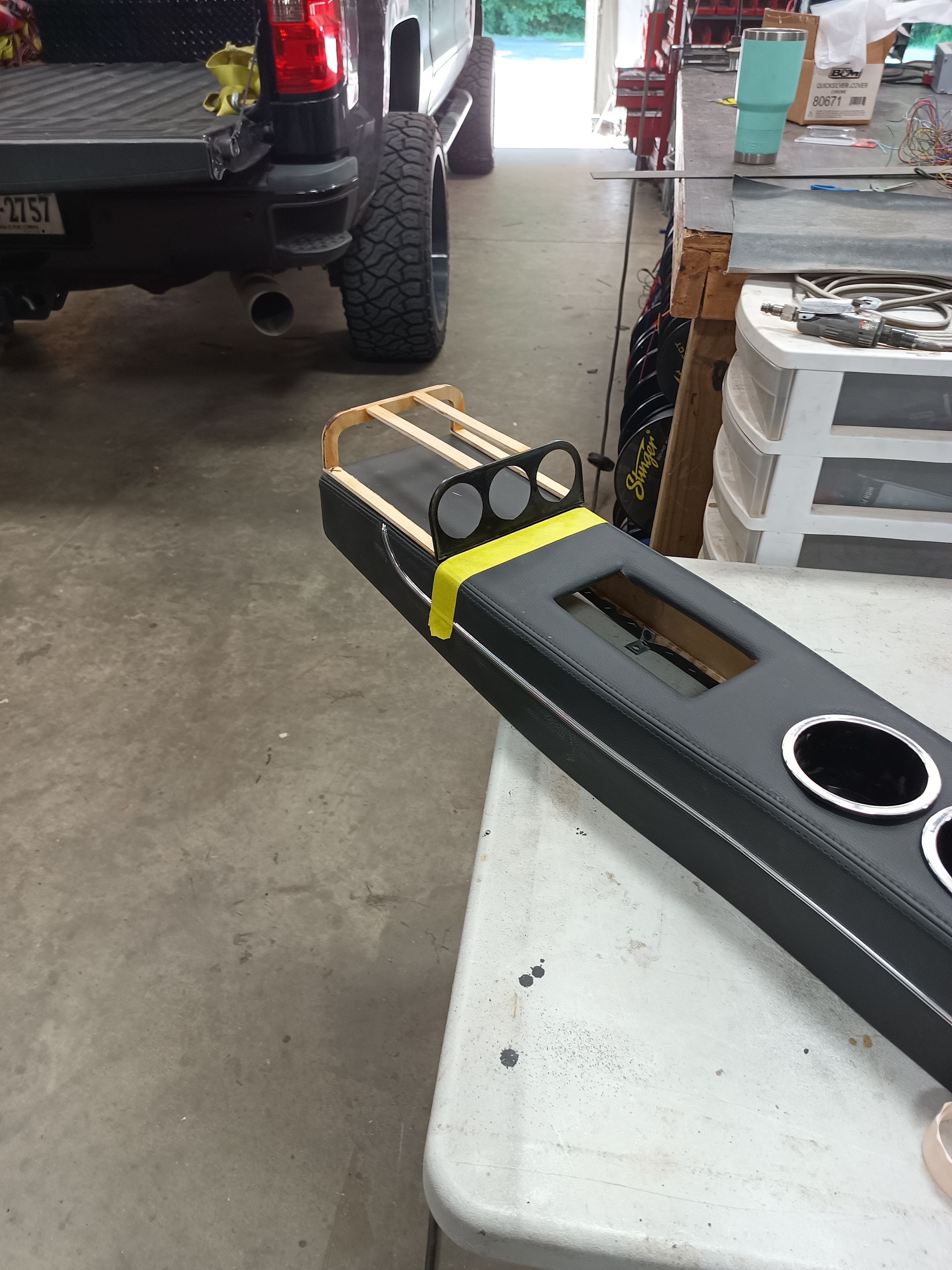 Black custom car console with gauges, cupholders, and stitching, on a workbench near a truck.