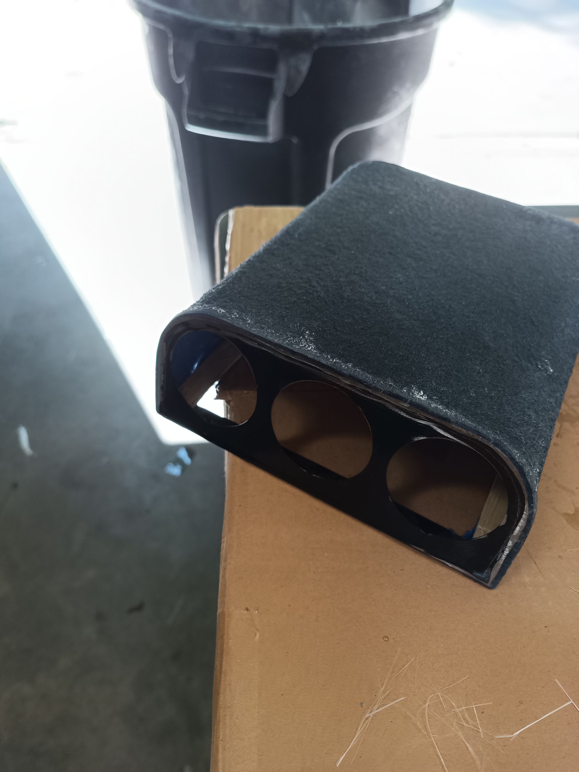 Black, rectangular object with three holes on a brown surface, next to a black trash can.