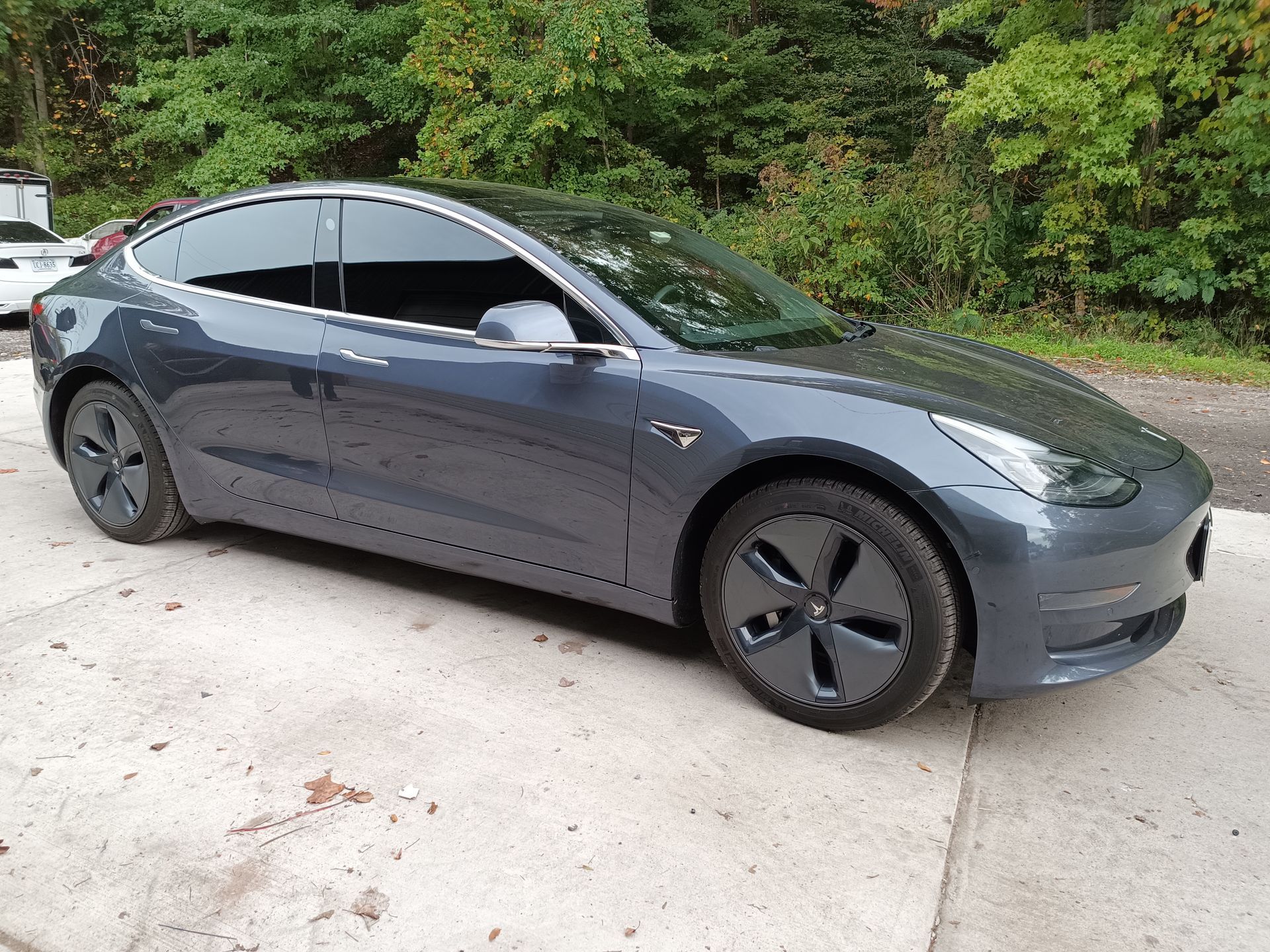 Gray Tesla Model 3 sedan parked on a concrete surface, with trees in the background.