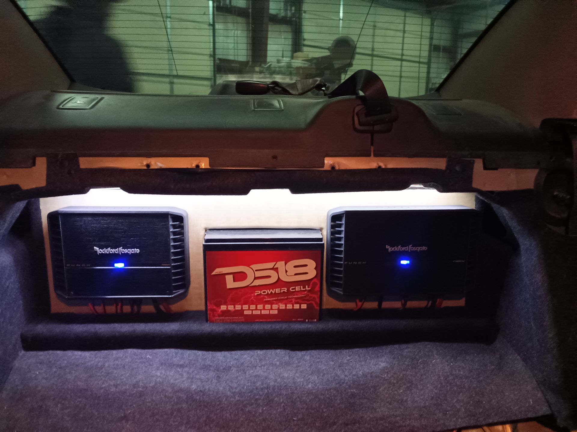 Car trunk with two black amplifiers and a red audio component; white LED strip.