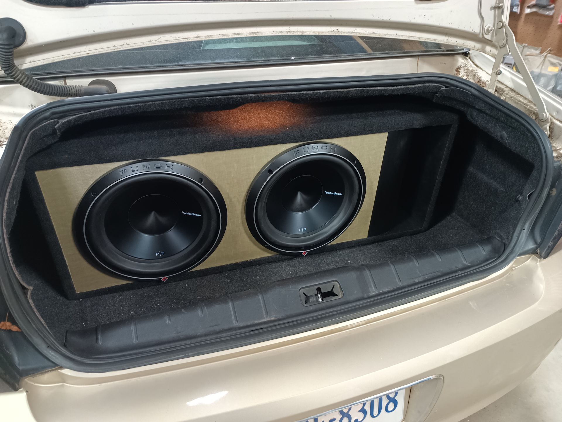 Two black subwoofers in a tan box mounted in the open trunk of a gold car.