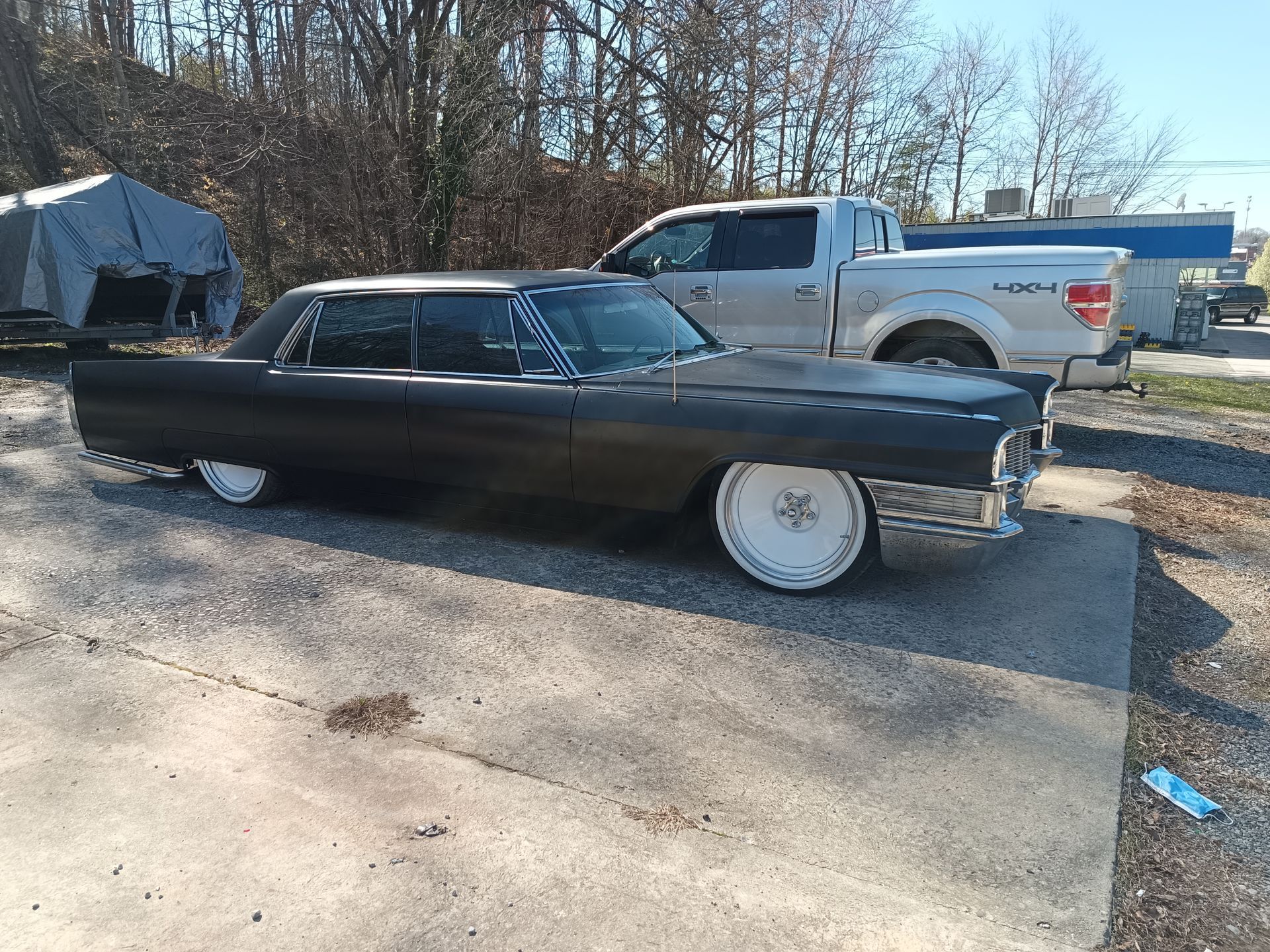 Black classic Cadillac with custom white rims parked outdoors near a truck and trees.