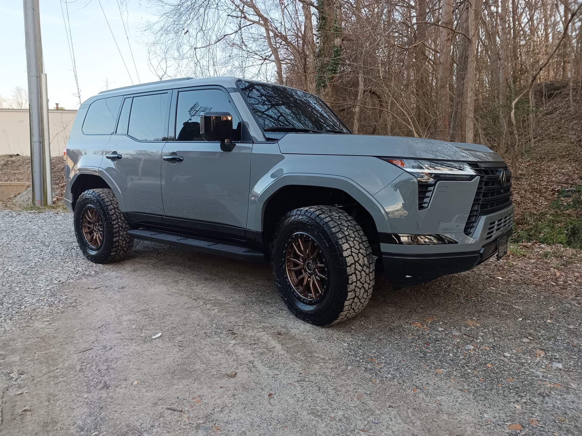 Gray SUV with bronze wheels parked on gravel. Daytime, wooded background.