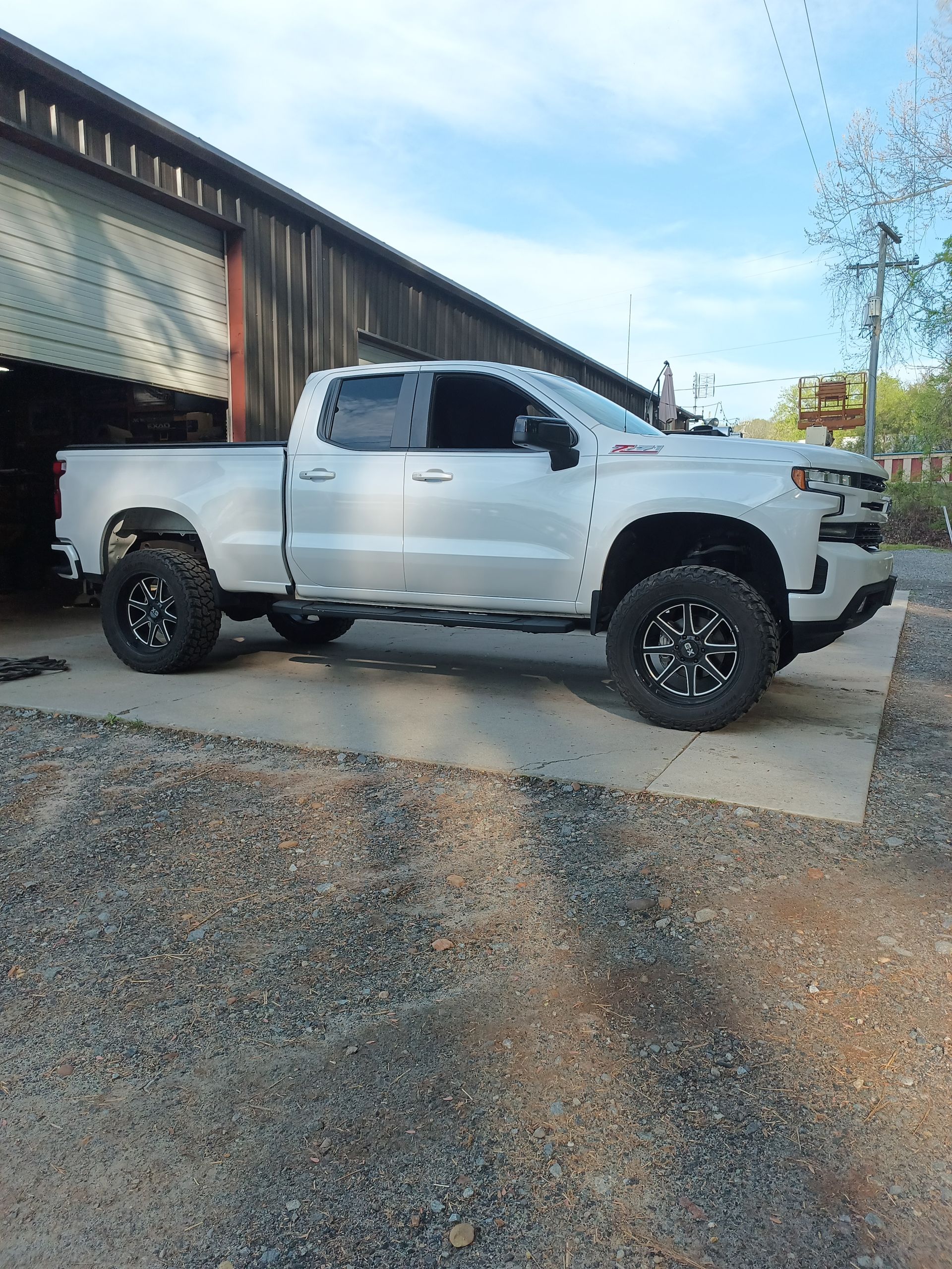 Silver Chevy pickup truck with black wheels parked on concrete. Gravel ground, cloudy sky.